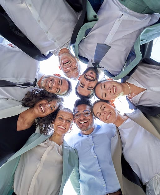 A low-angle, 'worm's-eye view' of a diverse group of eight smiling professionals in business-casual attire, standing in a tight circle and looking down into the camera against a bright, overexposed sky.